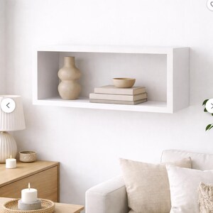 Op de afbeelding: Een witte rechthoekige zwevende plank met een beige vaas, een kleine kom en opgestapelde boeken. De plank is aan een witte muur bevestigd, wat een minimalistische en moderne esthetiek creëert. De items op de plank zijn neutraal van kleur.