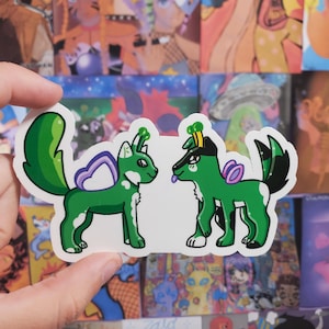 May include: A sticker featuring two green cartoon dog-like creatures with purple accents. The creatures have large tails and are facing each other. The background is filled with various colorful illustrations.