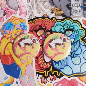 May include: Two yellow round buttons with a cartoon rat design, surrounded by colorful stickers. The rat is white and brown with pink accents, and the buttons have the word "FIG" on them. The stickers feature various cartoon characters and designs.