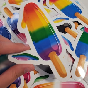 May include: A collection of colorful popsicle stickers. Each sticker features a popsicle design with a rainbow gradient of colors, including red, orange, yellow, green, blue, and purple. The popsicles have a wooden stick and a white border.