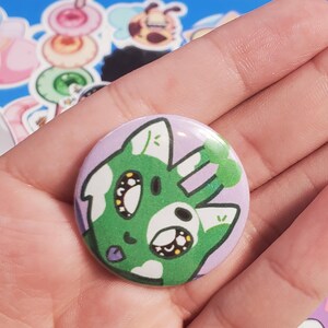 May include: A round button with a cartoon cat design. The cat has green fur with white spots and a purple background. The cat has large eyes and a small tongue. The button is held in a hand.