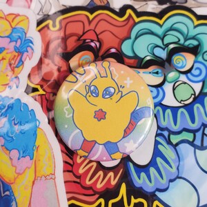 May include: A round, yellow button with a cartoon alien design. The alien has large blue eyes and a red star on its chest. The button is set against a background of colorful stickers with various designs and colors.