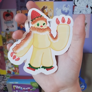 May include: A yellow and red cartoon character sticker with a strawberry on its head. The character has a white fluffy collar and red fingers. The sticker is on a hand.