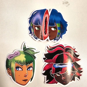 May include: Three die-cut stickers featuring stylized character heads. The top sticker shows a face split by a red fruit-like center with blue hair. The bottom left sticker has green hair and a purple geode-like accent. The bottom right has red and black hair with a blue geode-like accent.