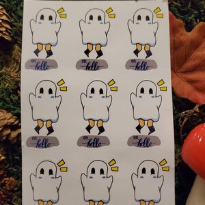 May include: Nine white ghost stickers with blue eyes and yellow accents. Each ghost is saying "Oh, hello" in blue text on a gray rock.