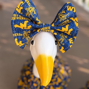 May include: A white goose figurine with a yellow beak wearing a large blue fabric bow featuring the West Virginia University logo and mascot in yellow. The fabric has a speckled blue pattern with repeating text and graphics of the university.