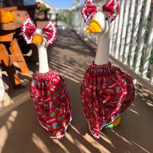 May include: Two white goose statues wearing red fabric outfits featuring an Ohio State Buckeyes pattern. Each goose has a matching red patterned bow attached to its head. The fabric has a white scalloped trim along the edges of the skirts and bows.