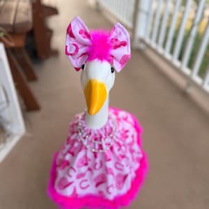 Pode incluir: Uma estátua de ganso branco vestindo uma roupa de tecido rosa e branco com estampa de chapéus de cowboy. A roupa inclui um laço combinando com um detalhe de pena rosa brilhante, uma bainha com babados e um colar de pérolas falsas.