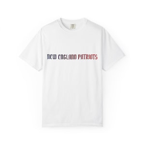 May include: A white short-sleeve t-shirt featuring the text NEW ENGLAND PATRIOTS printed across the chest in a gothic-style font. The word NEW is in dark blue, ENGLAND is in white, and PATRIOTS is in red, all centered on the front of the shirt.