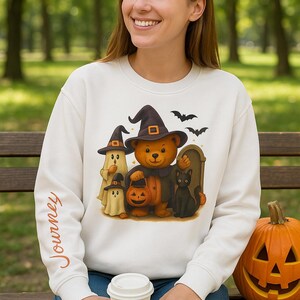 Puede incluir: Sudadera blanca con un diseño de Halloween que presenta un oso, fantasmas y un gato negro, todos con sombreros de bruja. Una calabaza tallada está en un banco. La palabra "Journey" está impresa en la manga.