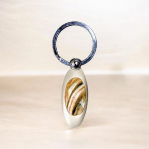 May include: A silver-colored keychain with an oval-shaped pendant. The pendant features a polished stone with brown, tan, and cream-colored swirling patterns. The keychain ring is circular and attached to the pendant with a small, silver-colored ball.