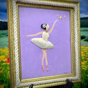 May include: A framed artwork featuring a ballerina in a white tutu, reaching for three gold stars. The artwork is set against a lavender background and framed in gold with a rope-like border. The artwork is displayed on a black easel.