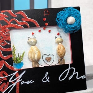 May include: A black picture frame with a red glitter design and the words "You & Me". The frame features two cat-shaped stone figures, a blue flower, and small red heart accents. A small blue pot with a plant is also visible.