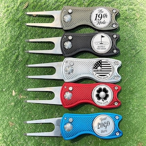 May include: Five metal golf divot repair tools with folding prongs and circular markers. The markers feature designs including The 19th Hole, a golf flag, an American flag, a four-leaf clover, and a blank space labeled Your Logo Here.