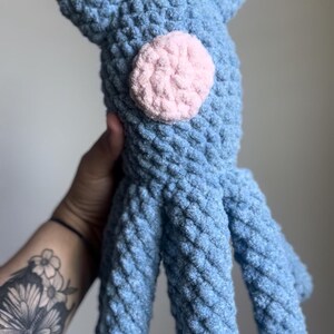 May include: A handmade blue crochet octopus plush toy with a pink circle accent. The plush has a textured, soft appearance and features eight tentacles. The toy is designed with two small ear-like protrusions on top.