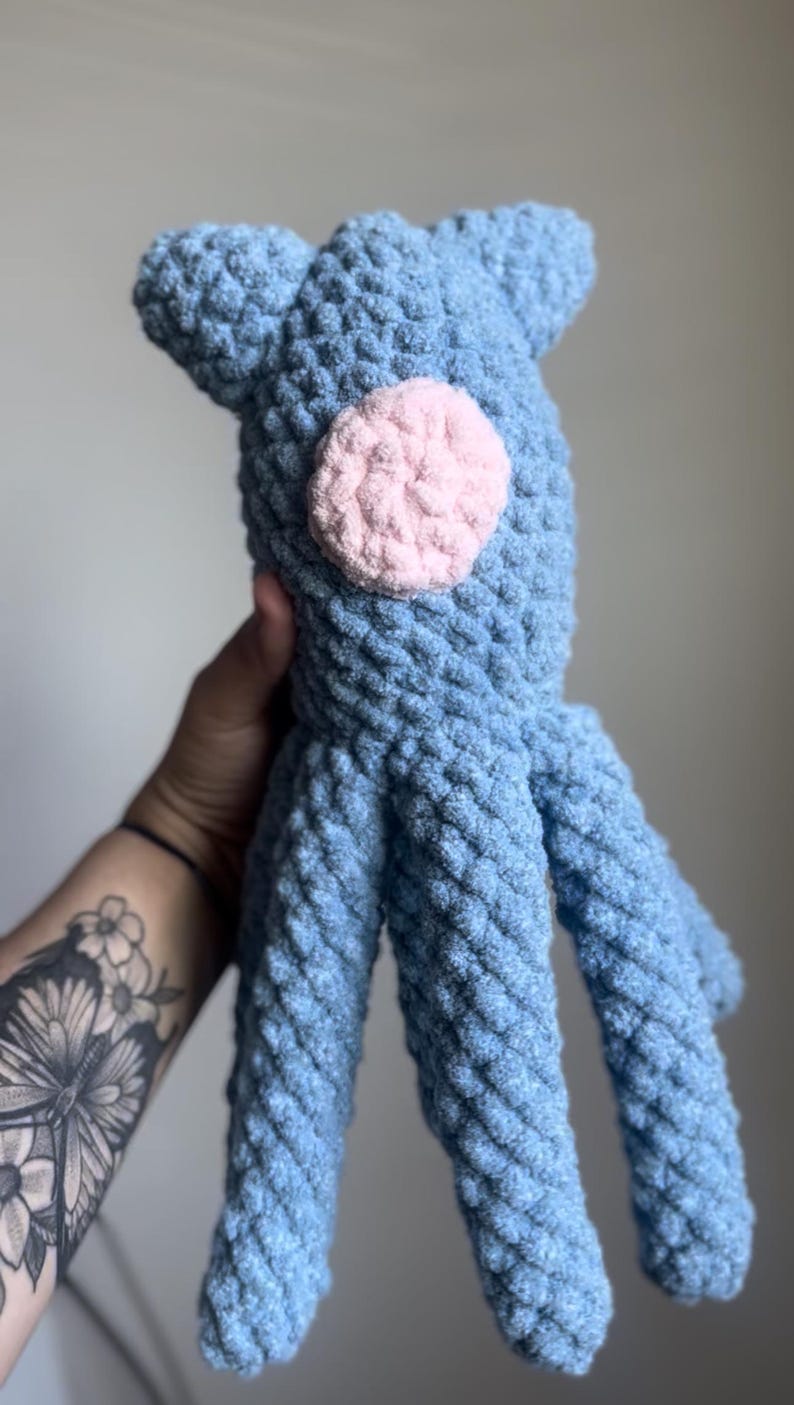May include: A handmade blue crochet octopus plush toy with a pink circle accent. The plush has a textured, soft appearance and features eight tentacles. The toy is designed with two small ear-like protrusions on top.