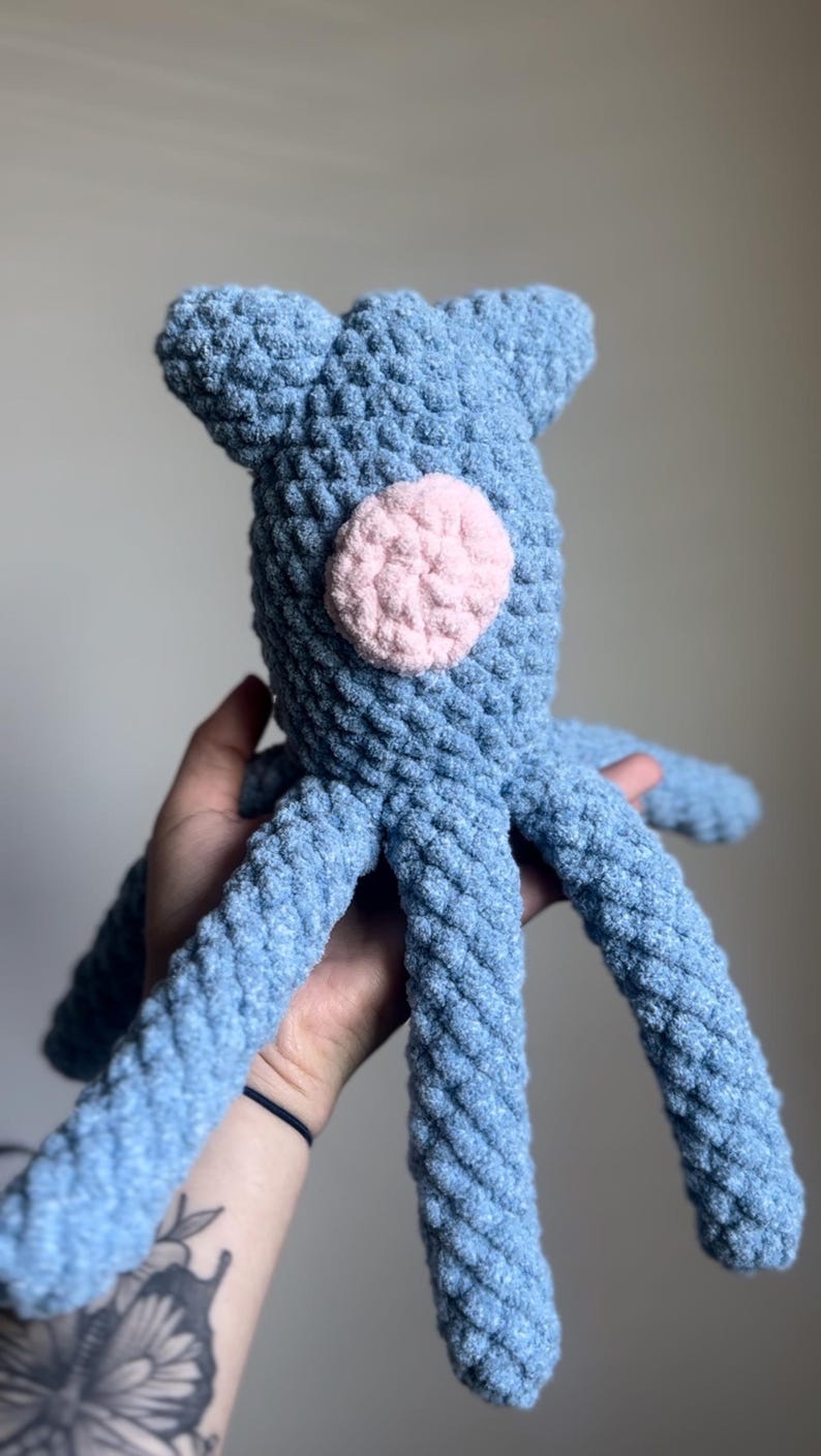 May include: A handmade blue crochet octopus with a pink circle on its body. The plush toy has a textured, soft appearance and is designed with eight tentacles. The octopus has two small ears on top of its head.
