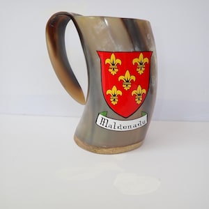 May include: A grey horn mug with a curved handle. It features a red shield with yellow fleur-de-lis symbols and the word "Maldonado" on a banner. The mug is displayed on a light surface. The mug is likely for drinking.
