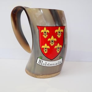 May include: A large, horn-shaped mug with a curved handle. The mug features a red shield with gold fleur-de-lis emblems and the name "Maldonado" on a banner. The mug is a mix of brown and gray.