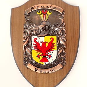 Może przedstawiać: Drewniana tarcza z miedzianym herbem. Herb przedstawia czerwonego orła na żółto-białej tarczy, z napisem "Fruzia" u góry i "France" na dole. Dekoracyjny przedmiot.