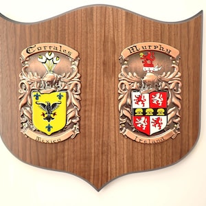 Może przedstawiać: Drewniana tablica z herbem rodzinnym z dwoma ozdobnymi metalowymi herbami. Lewy herb jest żółty z czarnym orłem i napisem "Corrales" powyżej. Prawy herb jest czerwono-biały z napisem "Murphy" powyżej. Tablica jest brązowa.