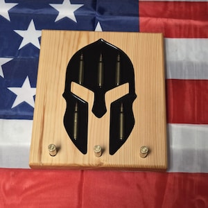 May include: Wooden wall decor featuring a black Spartan helmet design with bullet casings. The wooden plaque has three bullet casings inside the helmet and three on the bottom. The background is an American flag.