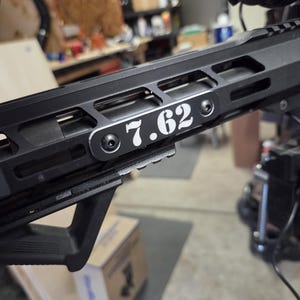 May include: Close-up of a black firearm with the text "7.62" in white. The firearm has a black handguard and a black angled foregrip. The background is a workshop setting.