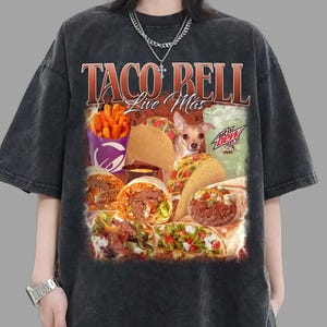 Puede incluir: Camiseta gris oscuro con un gráfico colorido que muestra comida de Taco Bell, incluyendo tacos y burritos. El diseño incluye el logotipo de Taco Bell y la frase "Live Mas". También aparece un perro pequeño.