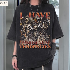 Puede incluir: Camiseta negra con aspecto desgastado, con un gr&aacute;fico de calaveras y el texto "I HAVE HOMOSEXUAL TENDENCIES" en naranja. El dise&ntilde;o incluye im&aacute;genes de calaveras sosteniendo armas y una parca.