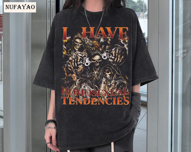 Puede incluir: Camiseta negra con aspecto desgastado, con un gr&aacute;fico de calaveras y el texto "I HAVE HOMOSEXUAL TENDENCIES" en naranja. El dise&ntilde;o incluye im&aacute;genes de calaveras sosteniendo armas y una parca.