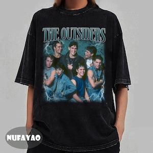 May include: Black t-shirt featuring a graphic of the movie "The Outsiders" with a group of actors. The title "THE OUTSIDERS" is printed above the image in a blue font. The shirt has a vintage, distressed look.