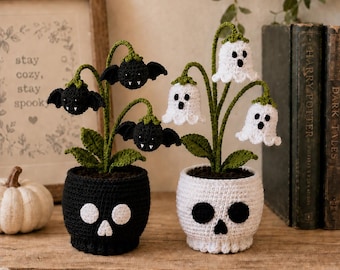 Puede incluir: Dos plantas de ganchillo en macetas con forma de calavera. Una maceta negra contiene tres flores negras con forma de murciélago y colmillos blancos. Una maceta blanca contiene tres flores blancas con forma de fantasma con ojos y bocas negros.