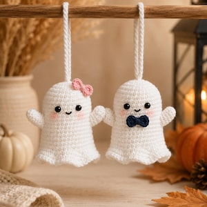 Pode incluir: Dois ornamentos de fantasma em crochê branco pendurados numa haste de madeira. O fantasma da esquerda tem um pequeno laço rosa, enquanto o da direita usa uma gravata borboleta azul-escura. Ambos têm olhos de contas pretas e bochechas rosadas.