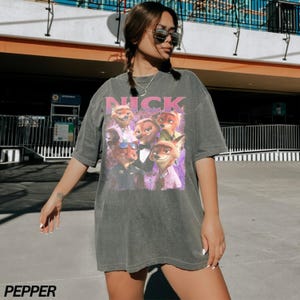 Op de afbeelding: Donkergrijze t-shirt met een afbeelding van het personage Nick Wilde uit de film Zootopia. Het woord "NICK" staat in roze boven de afbeelding. Het t-shirt heeft een vintage uitstraling.