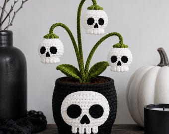 Puede incluir: Una planta tejida a ganchillo con tres flores blancas en forma de calavera con cuencas oculares y nasales negras sobre tallos verdes con hojas verdes, en una maceta negra tejida que muestra un gran motivo de calavera blanca en el frente.