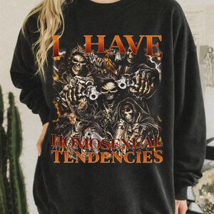 Puede incluir: Sudadera negra con el texto "I HAVE HOMOSEXUAL TENDENCIES" en naranja. El gr&aacute;fico presenta una ilustraci&oacute;n detallada de calaveras con armas y rel&aacute;mpagos.
