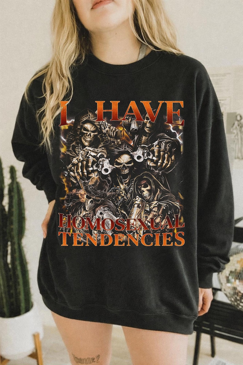 Puede incluir: Sudadera negra con el texto "I HAVE HOMOSEXUAL TENDENCIES" en naranja. El gr&aacute;fico presenta una ilustraci&oacute;n detallada de calaveras con armas y rel&aacute;mpagos.