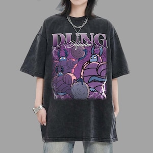Peut inclure: T-shirt gris foncé surdimensionné avec un motif de personnages de dessins animés violets et le texte "DUNG Defender" en violet. Le design comprend un effet d'éclair. Le t-shirt a des manches courtes et un col rond.