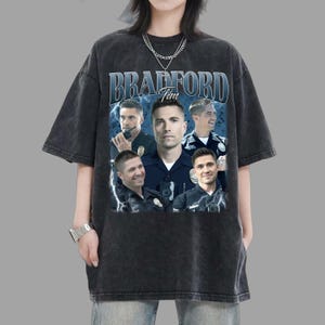 May include: Dark gray t-shirt featuring a collage of images and the text "BRADFORD Tim" in a blue and white design. The shirt has a vintage, washed appearance.