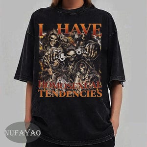 Puede incluir: Camiseta negra con texto naranja "I HAVE HOMOSEXUAL TENDENCIES". El dise&ntilde;o presenta un grupo de calaveras con armas y un fondo de fuego. La camiseta tiene mangas cortas y cuello redondo.