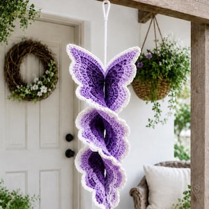 May include: A hanging spiral decoration crafted from purple crochet yarn with a white scalloped border. The structure features a series of connected butterfly-shaped segments that twist vertically, suspended by a white cord loop at the top.