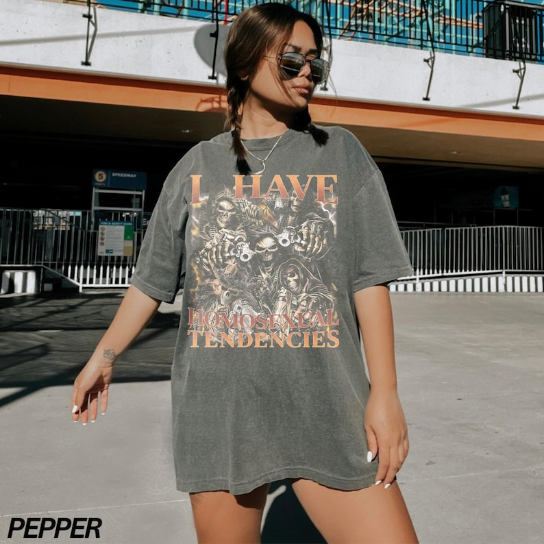 Puede incluir: Camiseta gris oscuro con el texto "I HAVE HOMOSOCIAL TENDENCIES" y un gr&aacute;fico de calaveras y figuras. La camiseta es de gran tama&ntilde;o y tiene mangas cortas.
