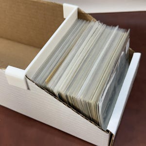 May include: A cardboard storage box with a white plastic divider, filled with a stack of cards. The box is open, revealing the contents. The cards are in individual protective sleeves.