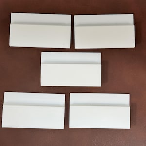 May include: Five white, rectangular, shelf-like objects with a lip, arranged on a brown surface. The objects appear to be made of a solid, opaque material. The lighting is even, and the objects are evenly spaced.