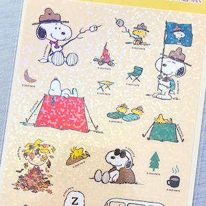 以下が含まれることがあります： スヌーピーとウッドストックが様々なキャンプシーンで描かれたピーナッツのステッカーシート。テント、キャンピングカー、寝袋、焚き火、木々のイラストがホログラム加工された背景にプリントされています。