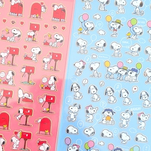 Może przedstawiać: Dwa arkusze naklejek z Snoopy z Peanuts. Jeden arkusz jest różowy z Snoopy w pobliżu czerwonej skrzynki pocztowej. Drugi jest niebieski z Snoopy trzymającym balony i noszącym kapelusze. Oba arkusze mają logo Peanuts i Hallmark.