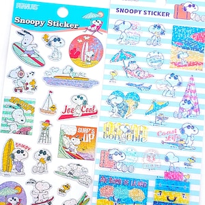 Könnte beinhalten: Zwei Blätter mit bunten Aufklebern, die Snoopy und andere Peanuts-Charaktere zeigen, die Sommeraktivitäten wie Surfen, Sonnenbaden und Entspannen am Strand genießen. Die Aufkleber befinden sich auf einem weißen Hintergrund mit blauen Streifen.