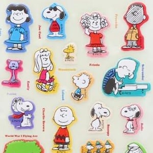 Peut inclure: Planche de stickers colorés avec divers personnages de Peanuts. Les autocollants incluent Snoopy, Charlie Brown, Lucy, et d'autres, avec le texte "Vintage Peanuts Stickers" et "Happiness is 75 Years of Peanuts".