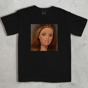 Może przedstawiać: Czarny t-shirt z kwadratowym zdjęciem twarzy lalki wyrażającej zaskoczenie. Lalka ma rude włosy, duże oczy i ściągnięte usta. T-shirt jest wykonany z miękkiego materiału.