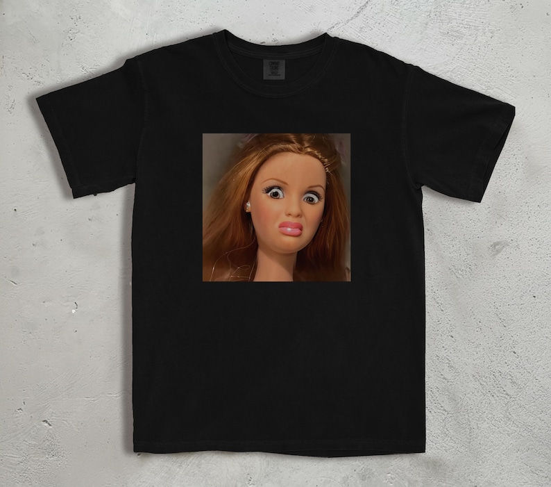 Może przedstawiać: Czarny t-shirt z kwadratowym zdjęciem twarzy lalki wyrażającej zaskoczenie. Lalka ma rude włosy, duże oczy i ściągnięte usta. T-shirt jest wykonany z miękkiego materiału.